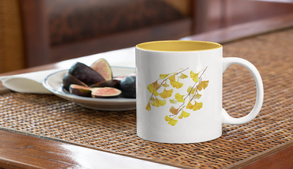 White mug with yellow interior and leaf design on a wooden table with figs in the background